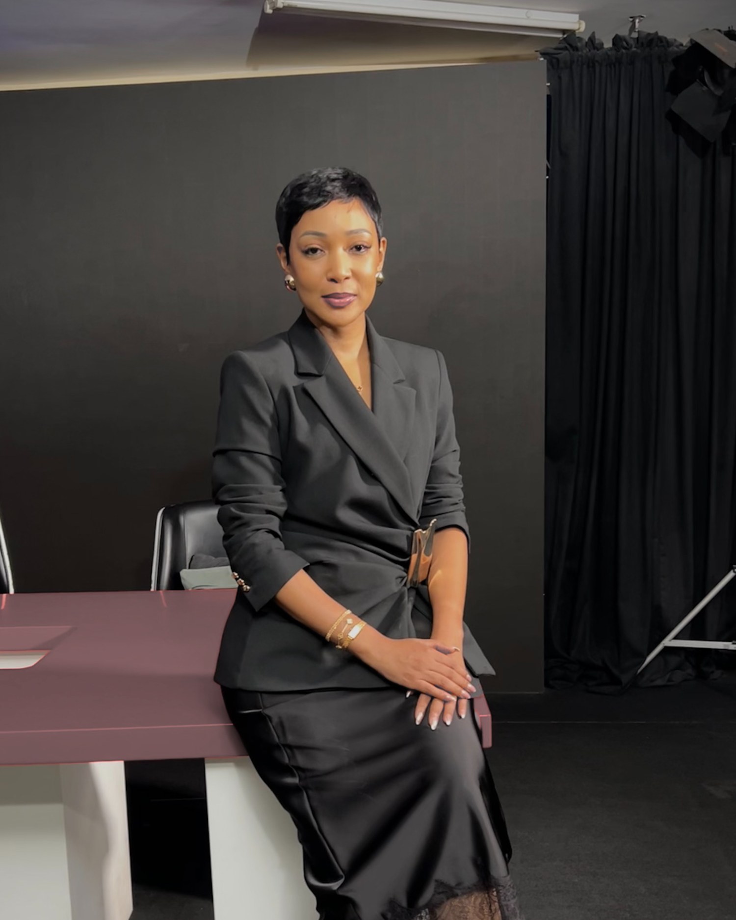 Yolanda Robert in a black tailored outfit, seated at the edge of a studio desk — editorial service portrait